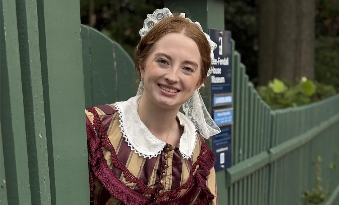 A smiling woman in historical dress posing by a green fence at Lee-Fendall House Museum.