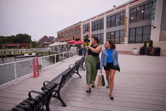 Two women carrying shopping bags walk along the Alexandria waterfront path next to a building.