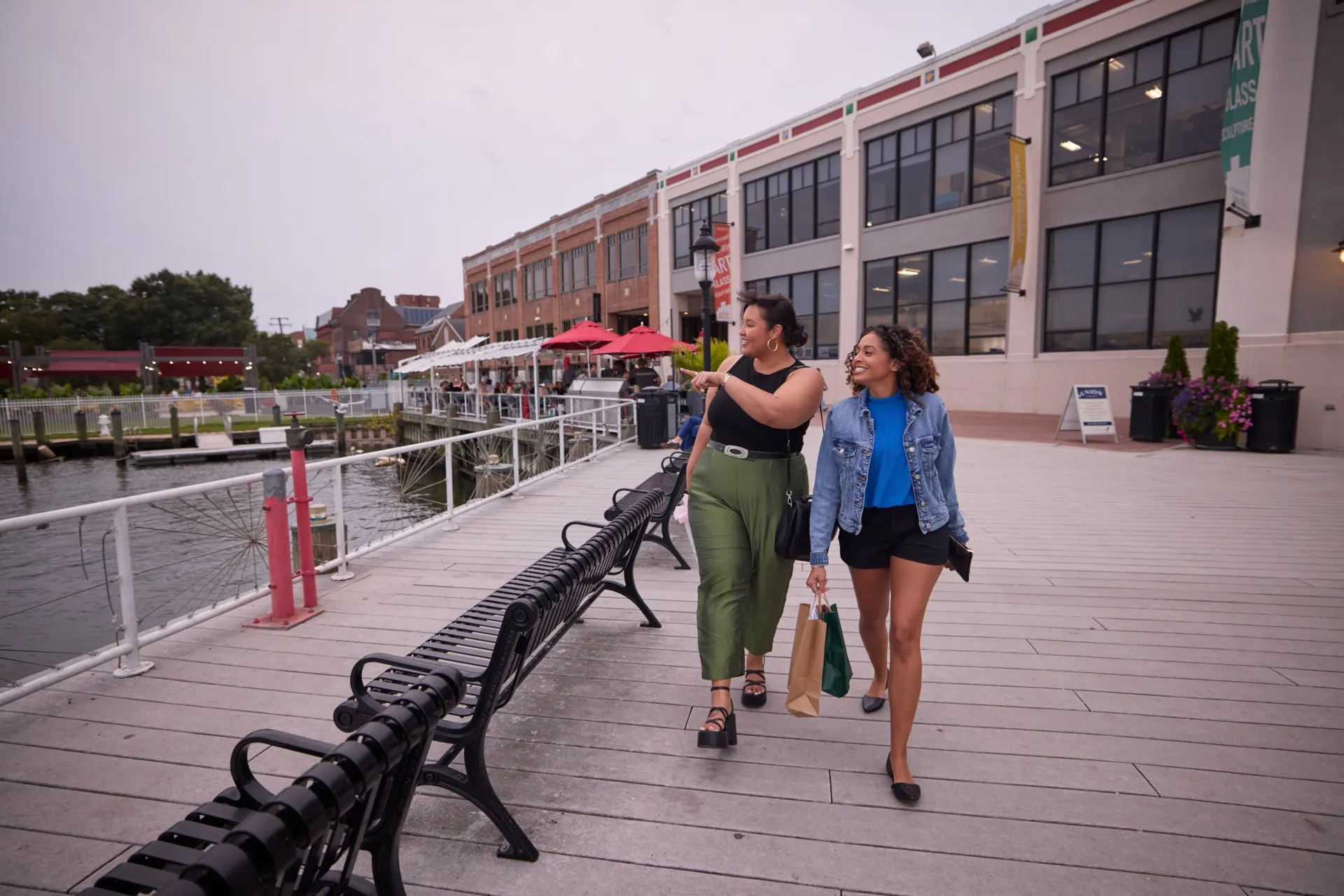 Two women carrying shopping bags walk along the Alexandria waterfront path next to a building.