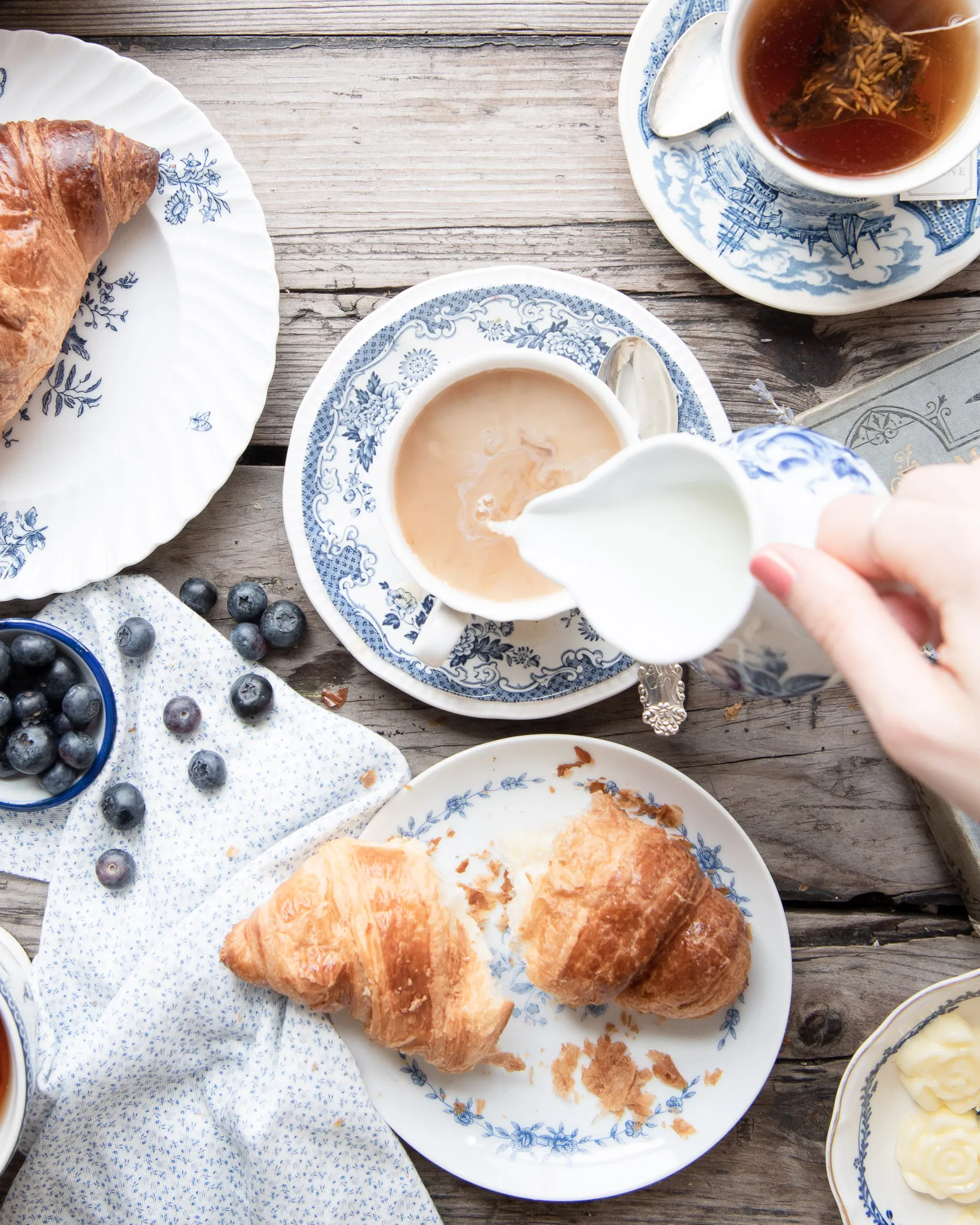 A close-up, top-down view of a hand pouring milk from a white pitcher into a cup of tea, surrounded by croissants and fresh blueberries.