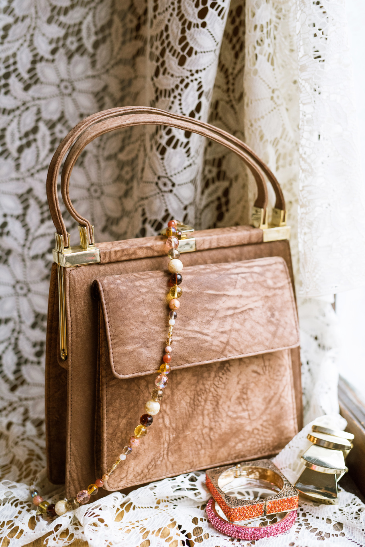 An elegant brown leather purse on a intricate lace curtain alongside other vintage items like bracelets, and beaded necklaces. 