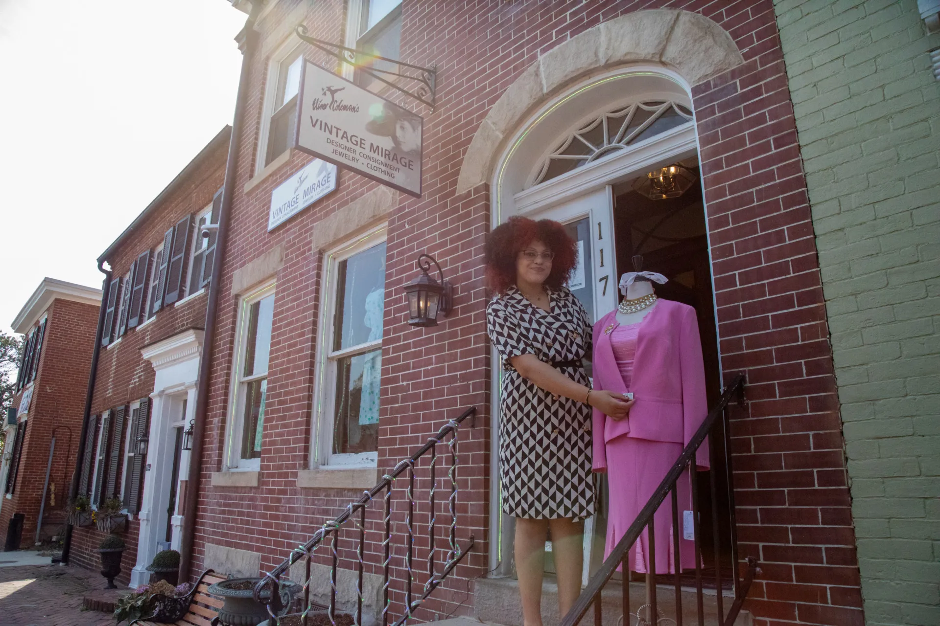 Exterior shot of Vintage Mirage's red brick building, general manager Rebecca Jacobs, is shown standing next to a mannequin display wearing a bright pink dress.
