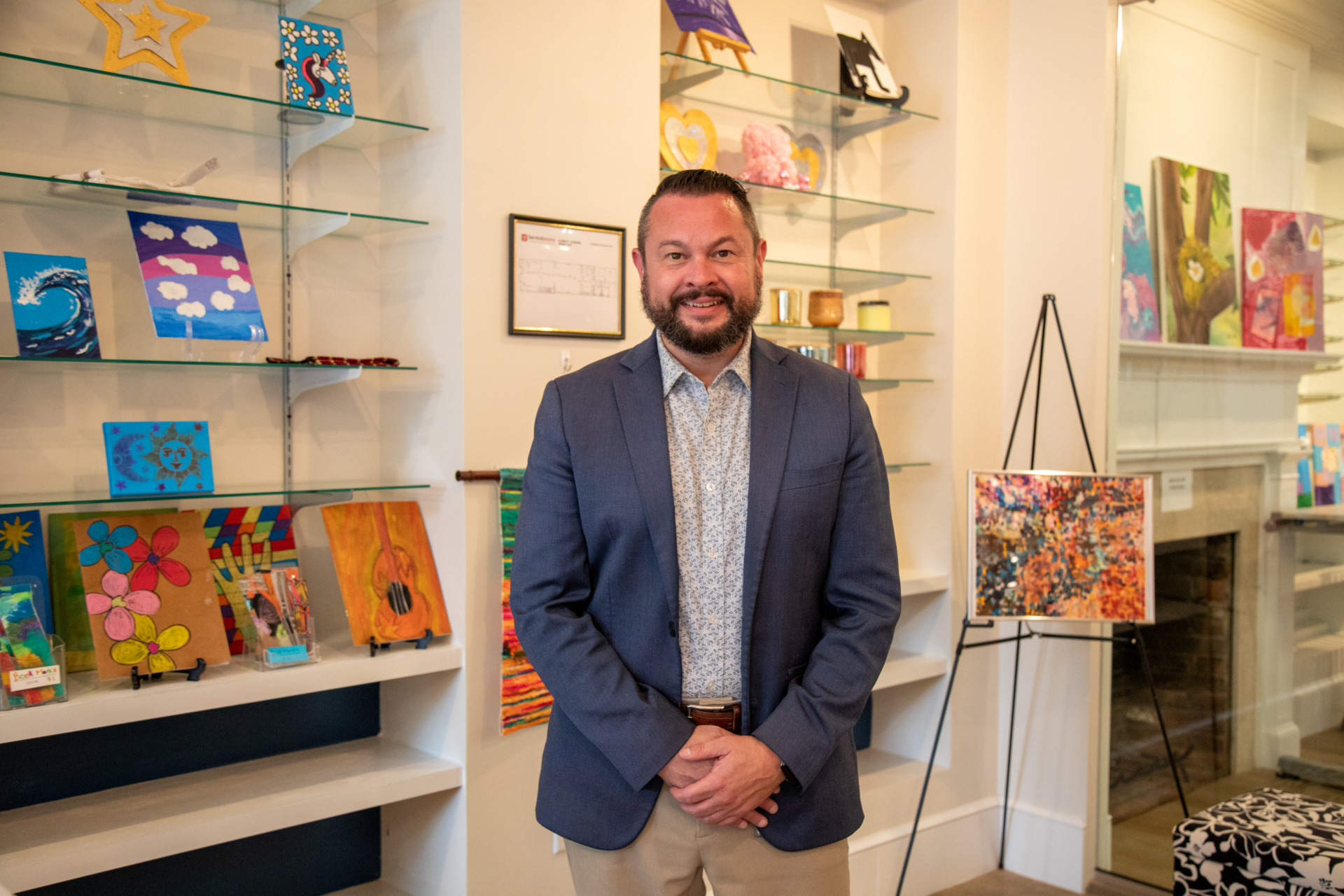 Joel Pace stands in front of a display of artwork including fabric handbags, abstract paintings, and more.