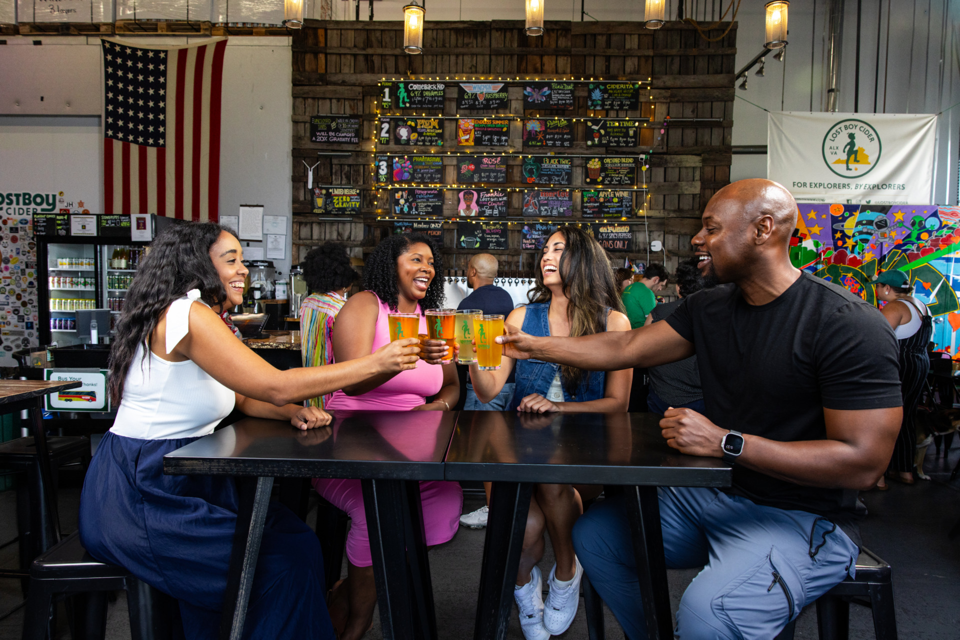 A group of people seated at a table at Lost Boy Cider, cheering with glasses of cider in hand, a colorful chalkboard menu behind the group.