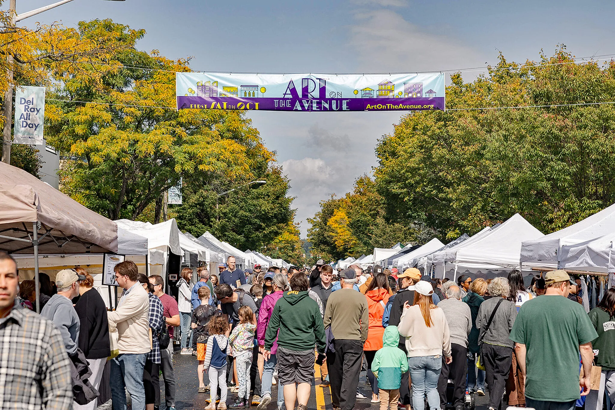 A row of tents lined along Mount Vernon Avenue in Del Ray. Pedestrians are shown walking down the street during Art on the Avenue. The festivals banner is hung above them.