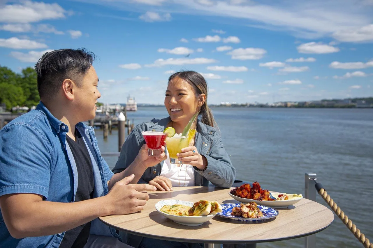 Two people clink their cocktail glasses at a high top table overlooking the Potomac River on a sunny summer day.