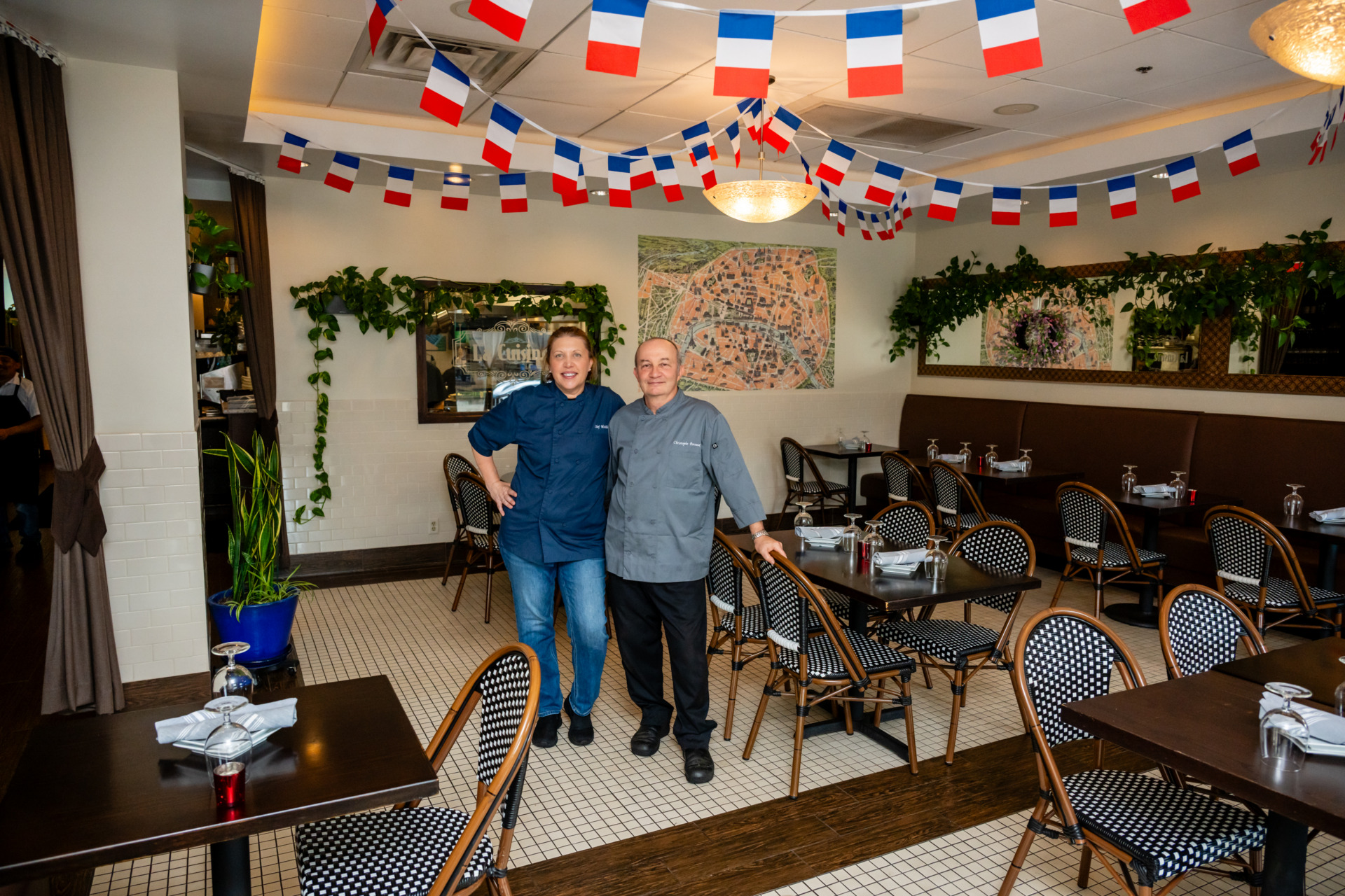 Chefs Michelle and Christophe standing inside the french-inspired dining room at Bastille. France's flags are hung on the ceiling. A map of France is hung on the wall, and Parisian-inspired furniture decorate the room.